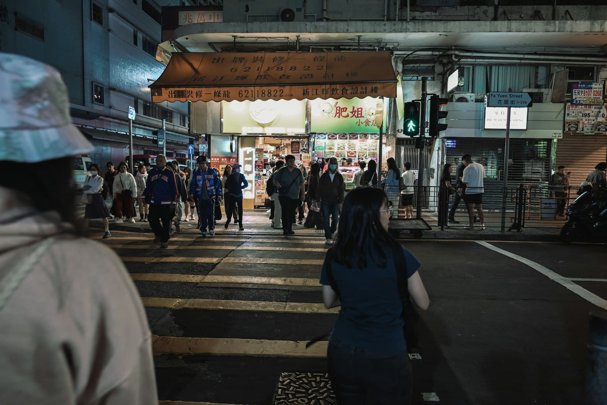 A crowd crossing a Hong Kong street towards one brightly lit shopfront, a figure out of focus in the foreground