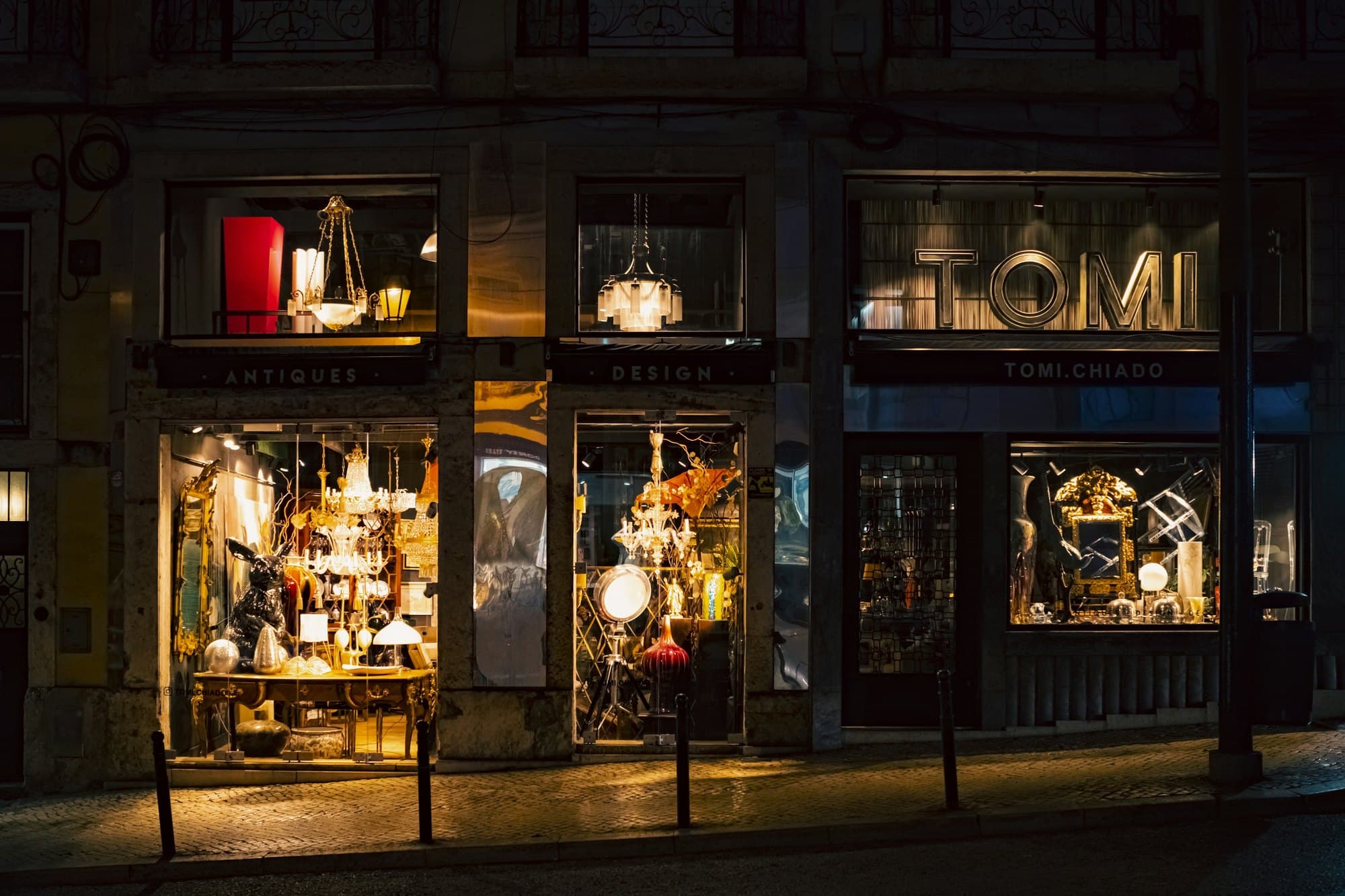 An antiques and design shop in Lisbon at night, its windows lit and its stock visible from the pavement