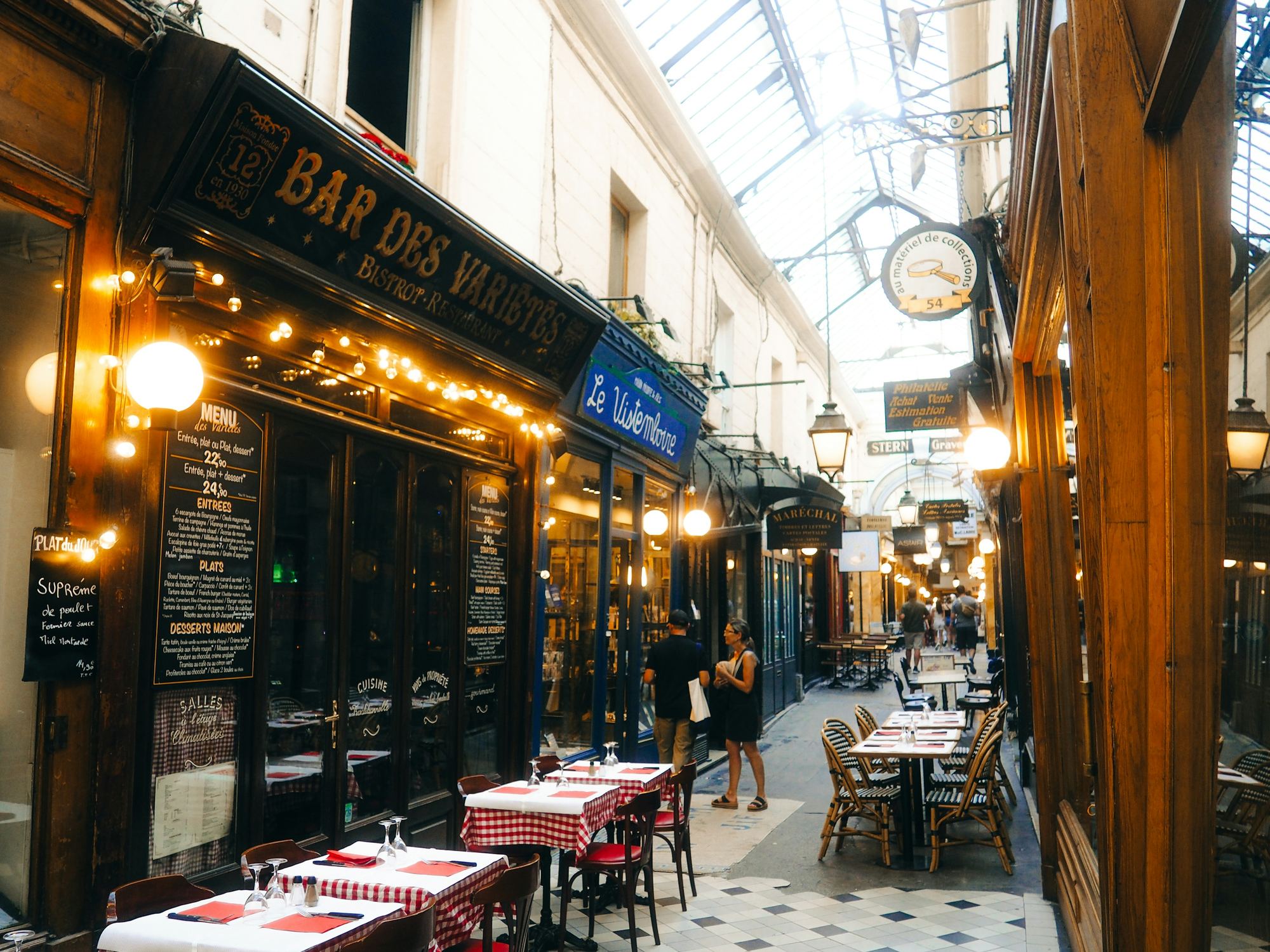 A glass-roofed Paris passage lined with bistro tables, lamps and shopfronts running into the distance
