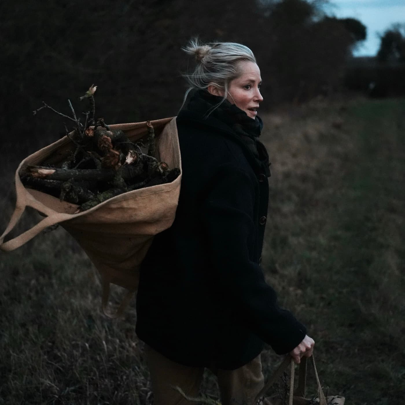 A woman carrying a bundle of kindling across a Norfolk field in a Carrier Company coat