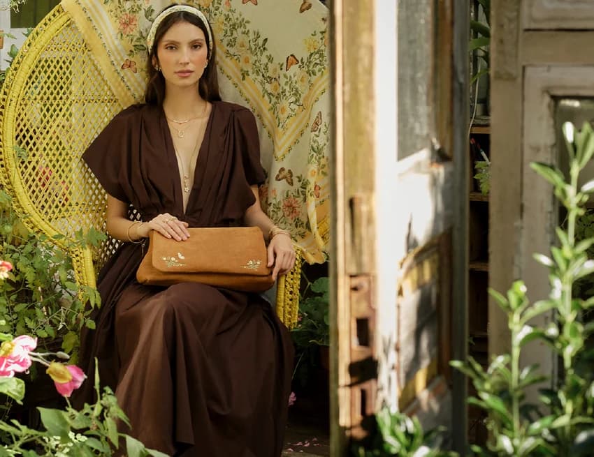 A model in a Fable England brown dress holding an embroidered clutch bag, seated in a peacock chair