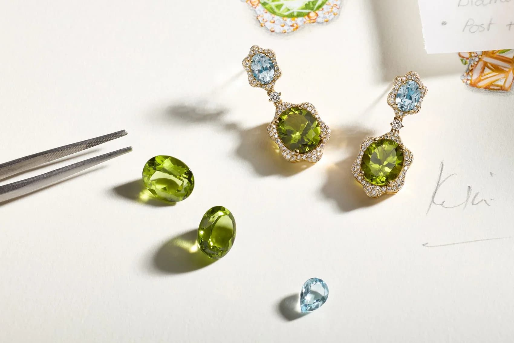 Kiki McDonough peridot and aquamarine drop earrings beside loose gemstones and a design sketch