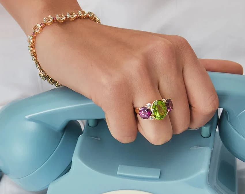 A hand wearing a Kiki McDonough peridot and tourmaline ring and bracelet, resting on a vintage telephone