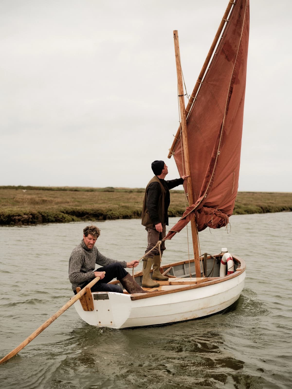 Two men sailing a wooden dinghy off the saltmarsh, both in Le Chameau wellington boots