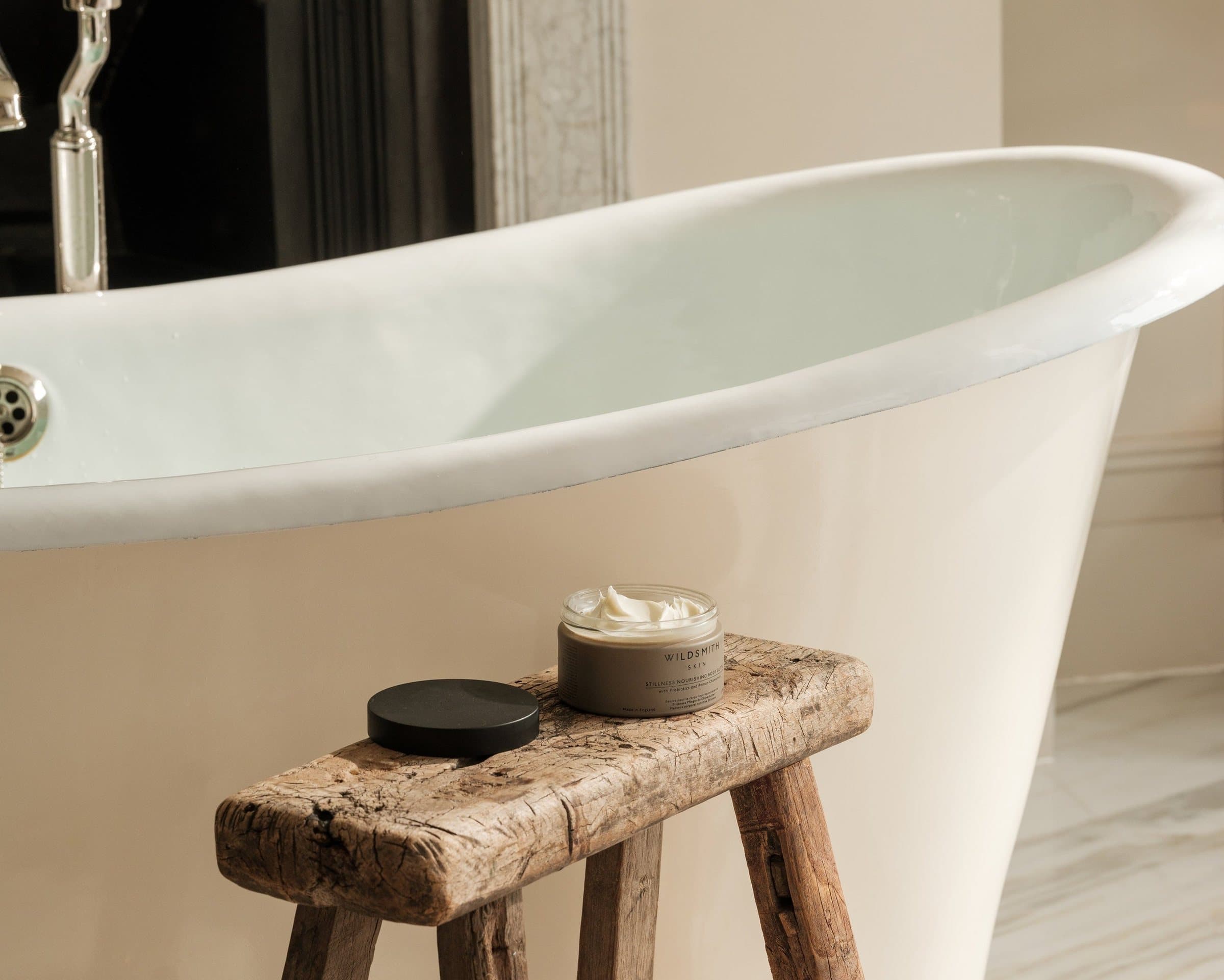 A jar of Wildsmith Skin cream on a weathered wooden stool beside a roll-top bath