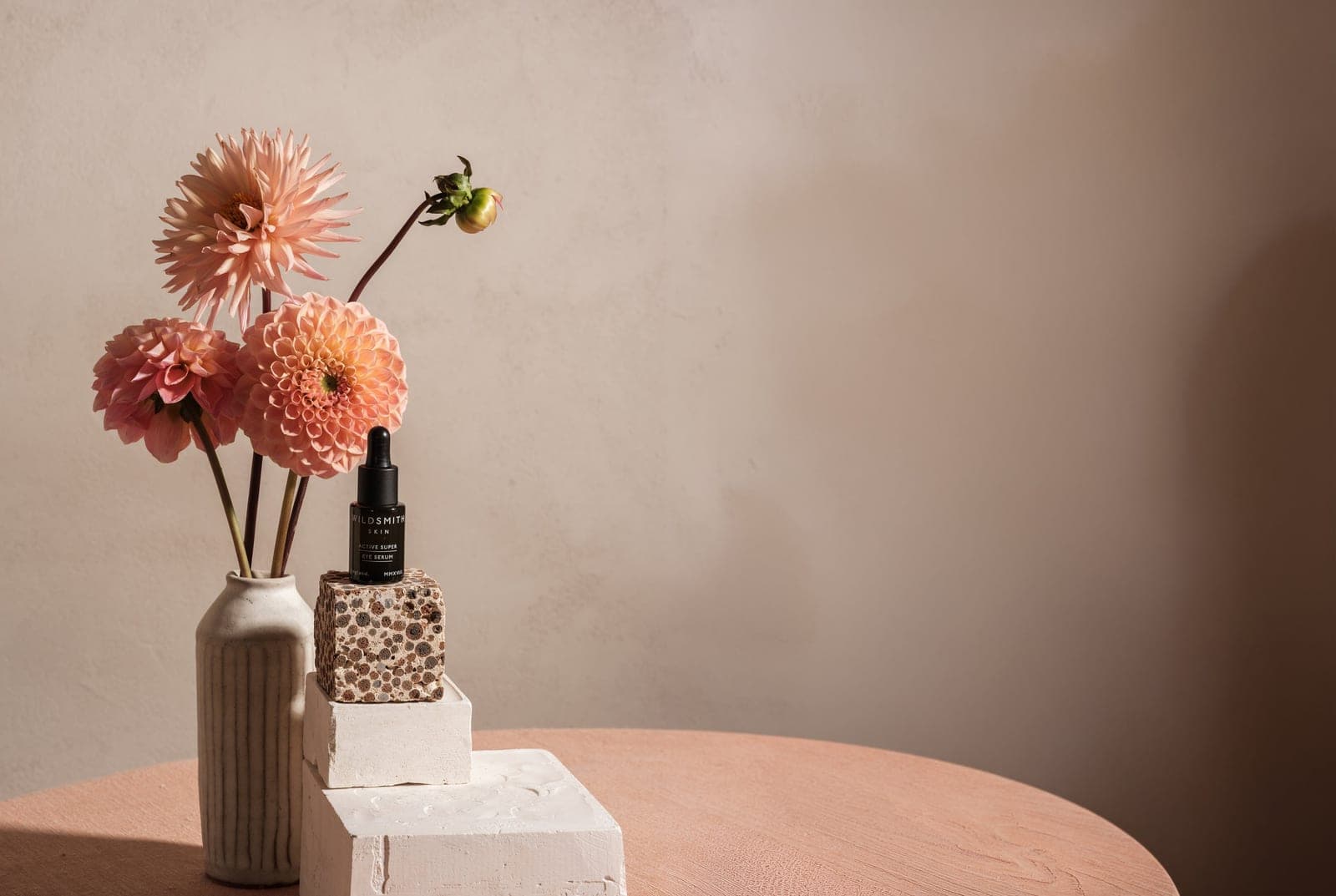 Wildsmith Skin Active Super Eye Serum beside a vase of dahlias, on a plaster plinth