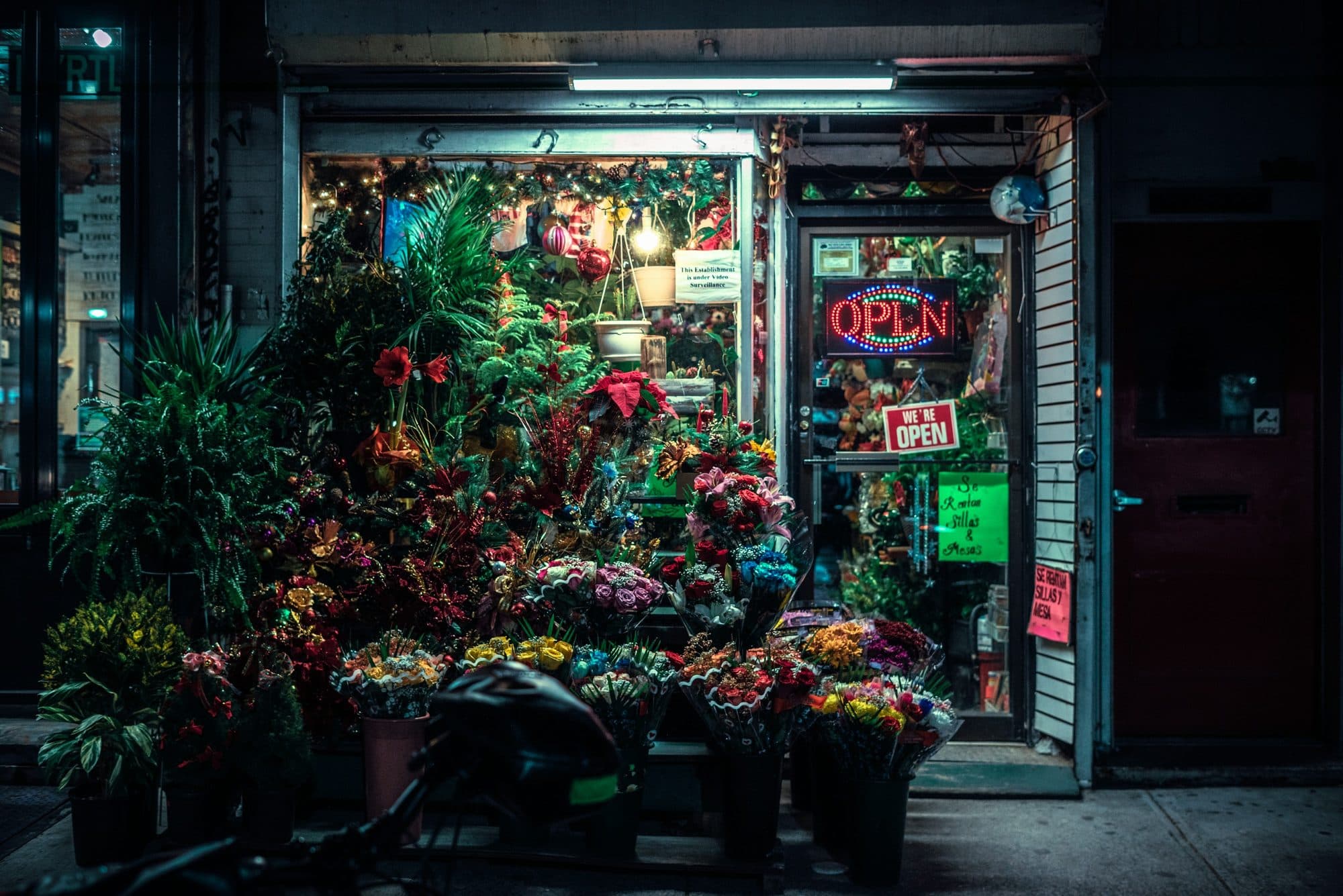 A florist trading late, buckets of cut flowers spilling onto the pavement under a neon Open sign