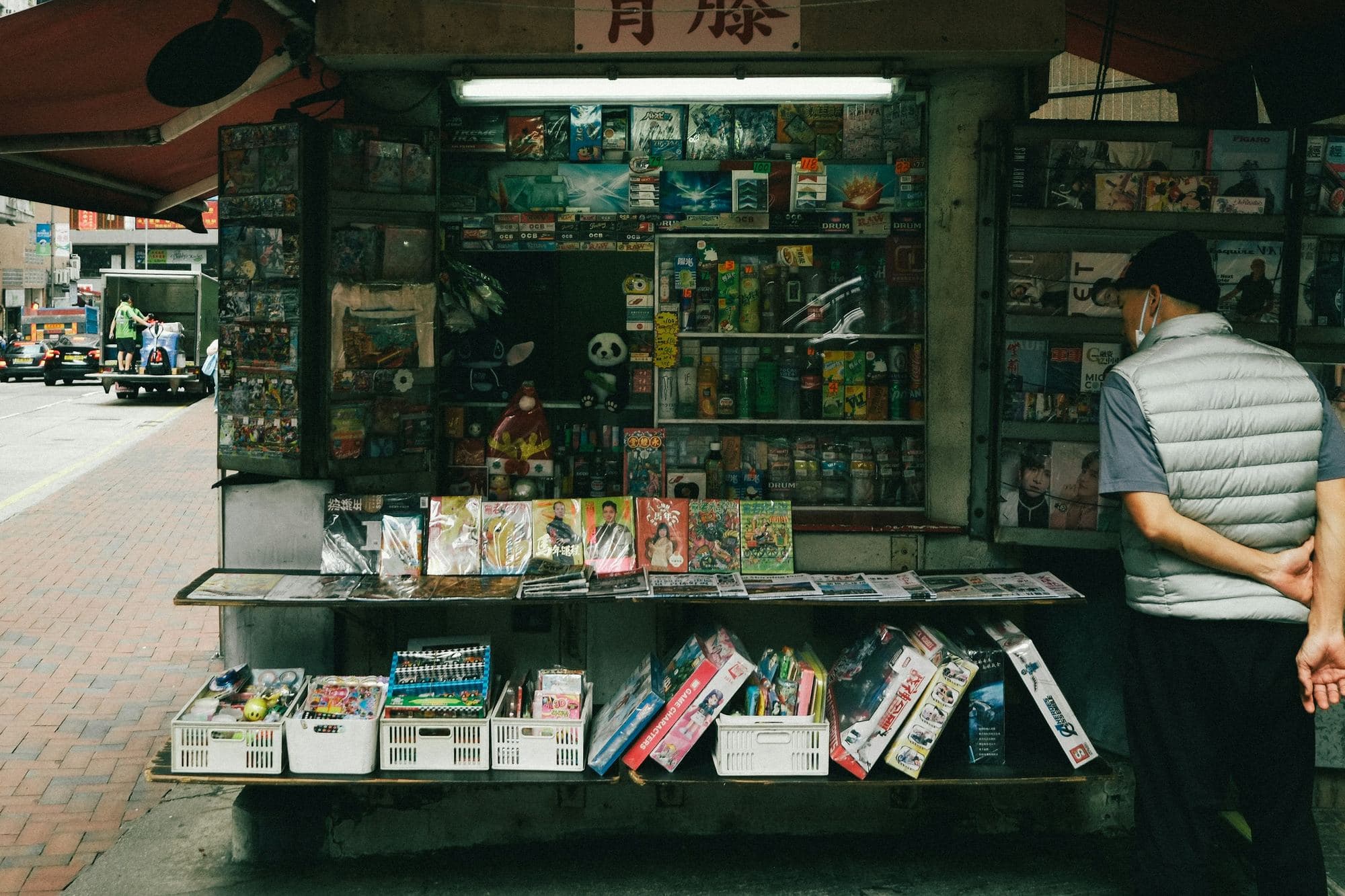 A man standing at a street kiosk, hands behind his back, reading the covers on display