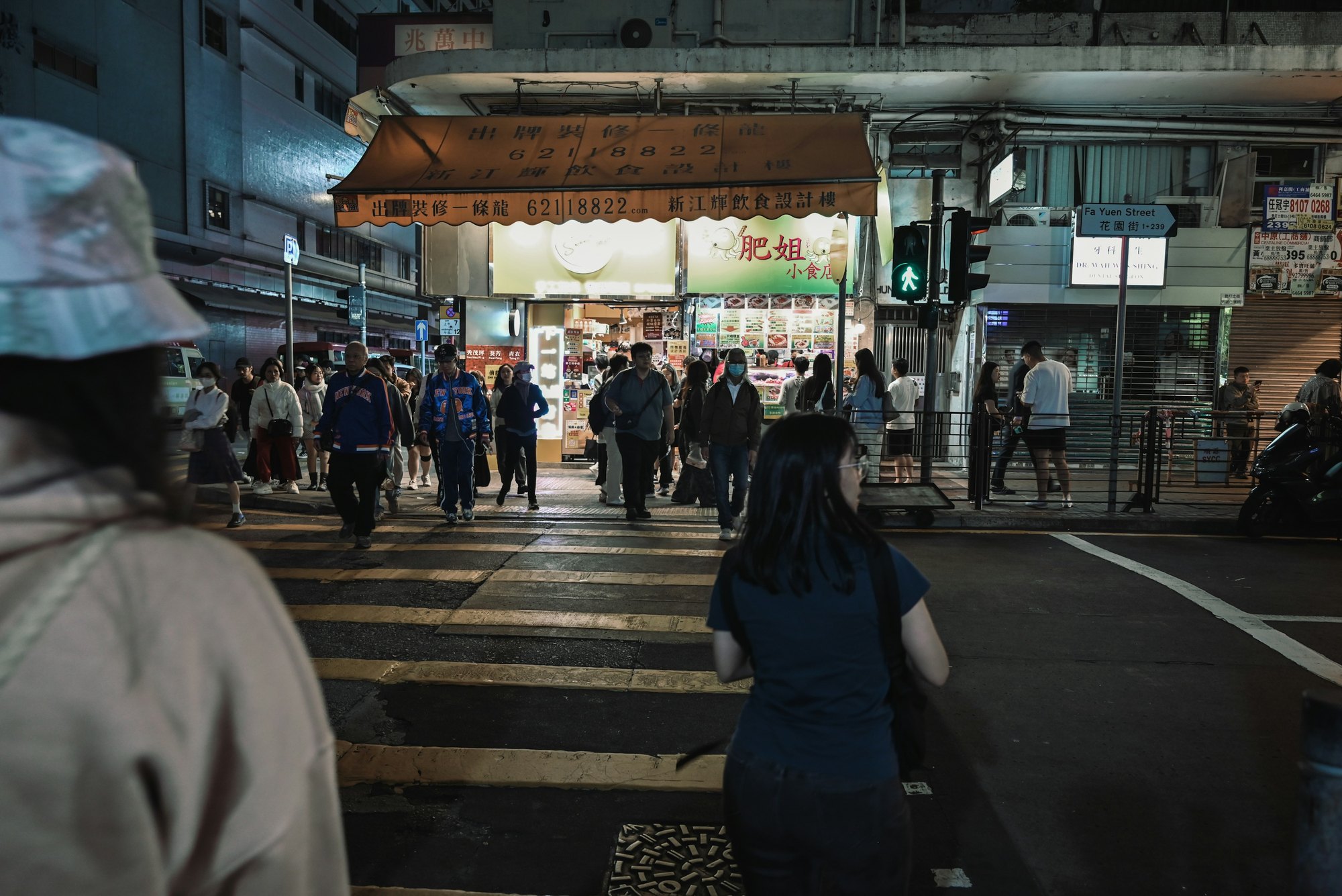 A crowd crossing a Hong Kong street towards one brightly lit shopfront, a figure out of focus in the foreground