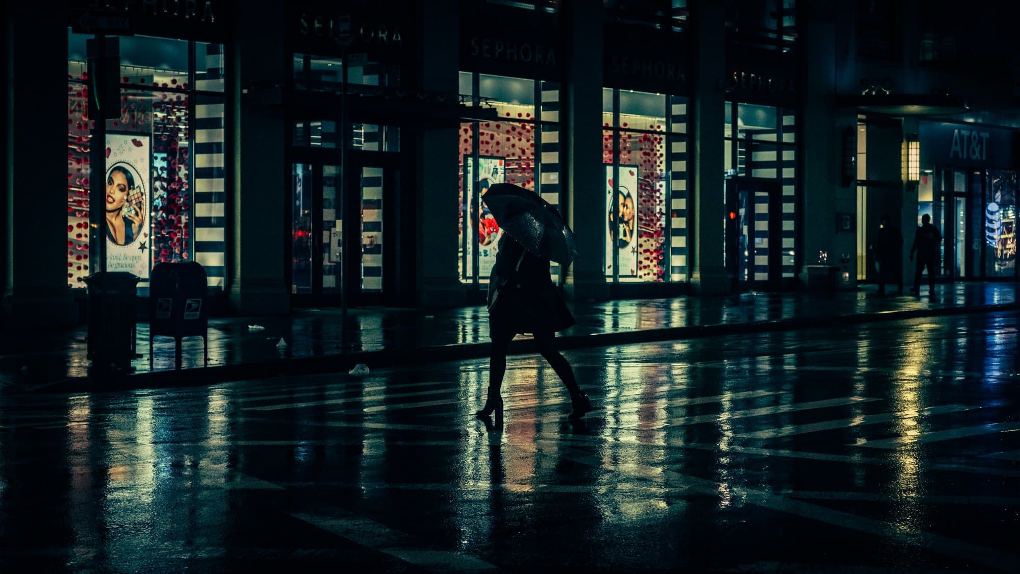 A lone figure with an umbrella crossing a wet street at night, lit shop windows reflected in the road