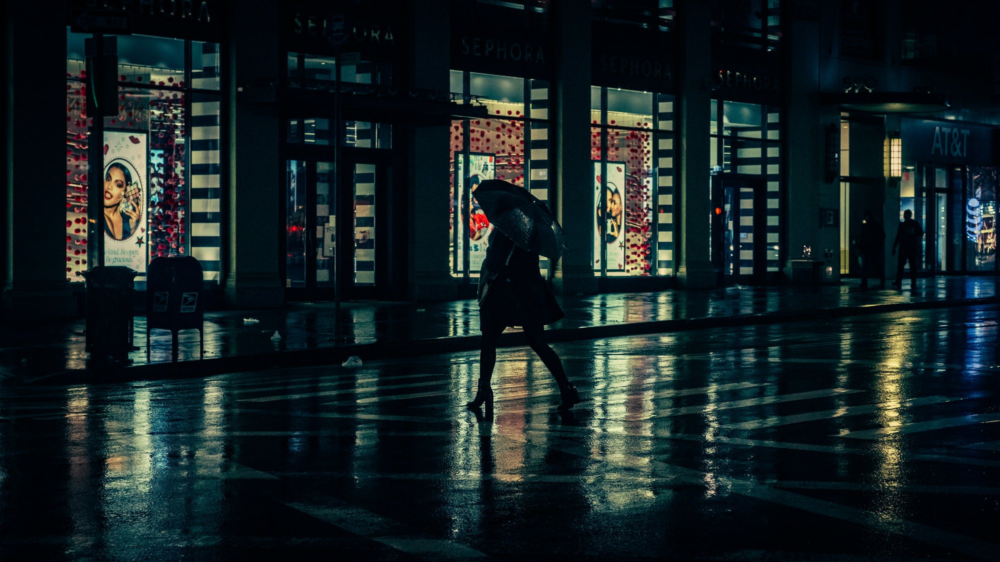 A lone figure with an umbrella crossing a wet street at night, lit shop windows reflected in the road