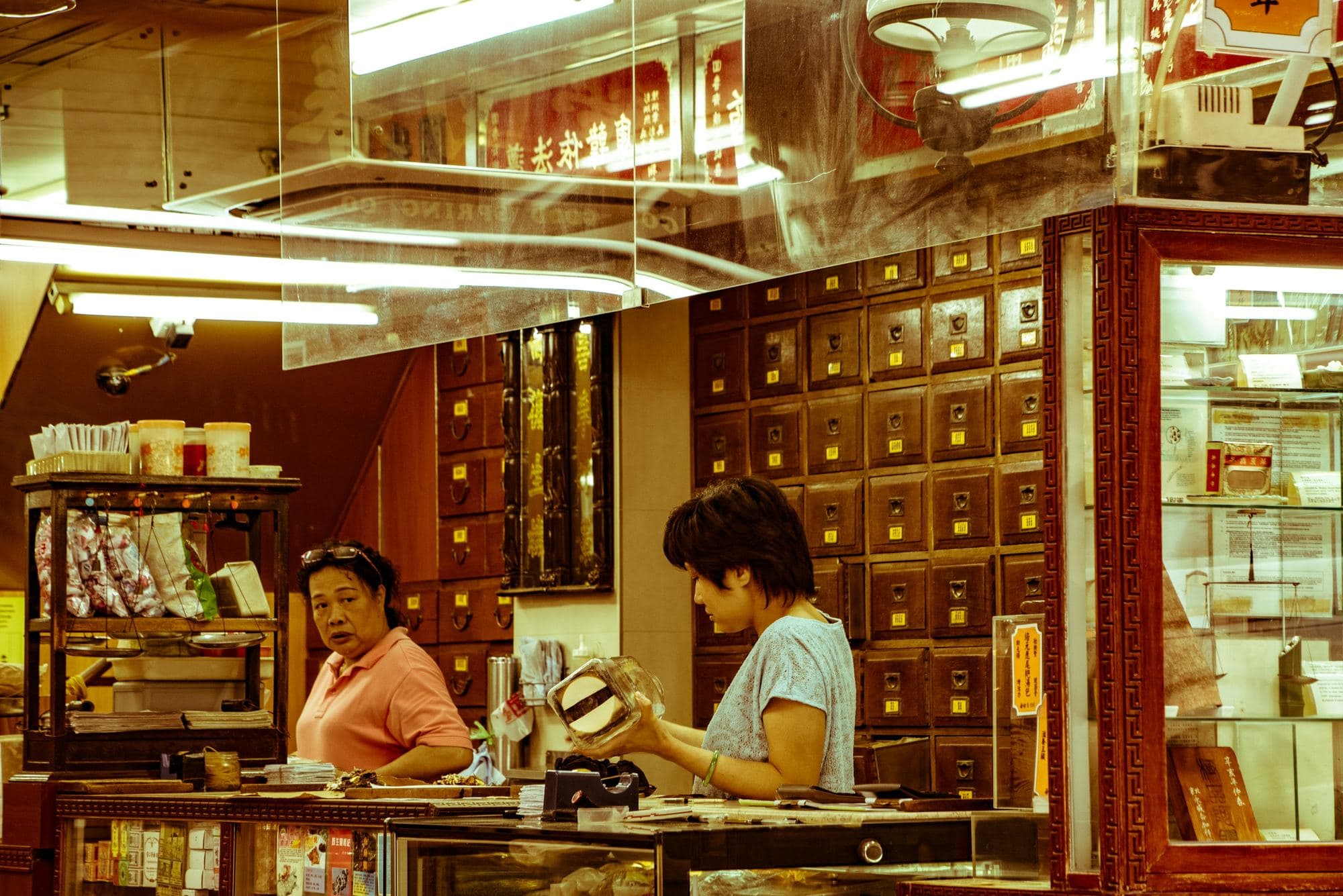 Two women working the counter of a Chinese medicine shop, a wall of labelled drawers behind them