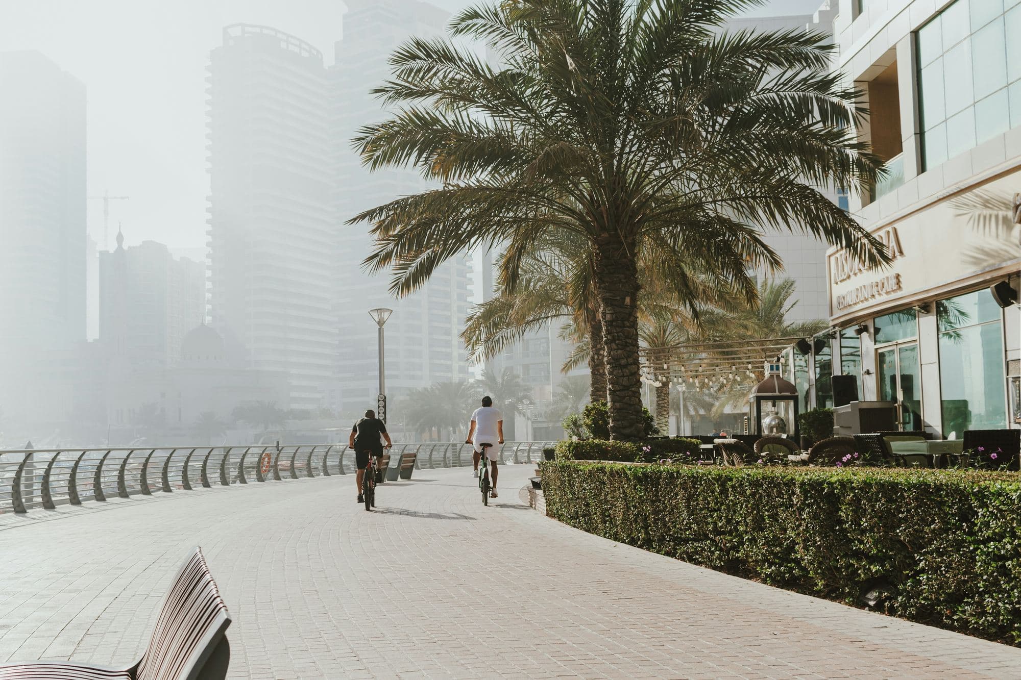 Two cyclists on a Dubai promenade, the towers behind them fading into morning haze