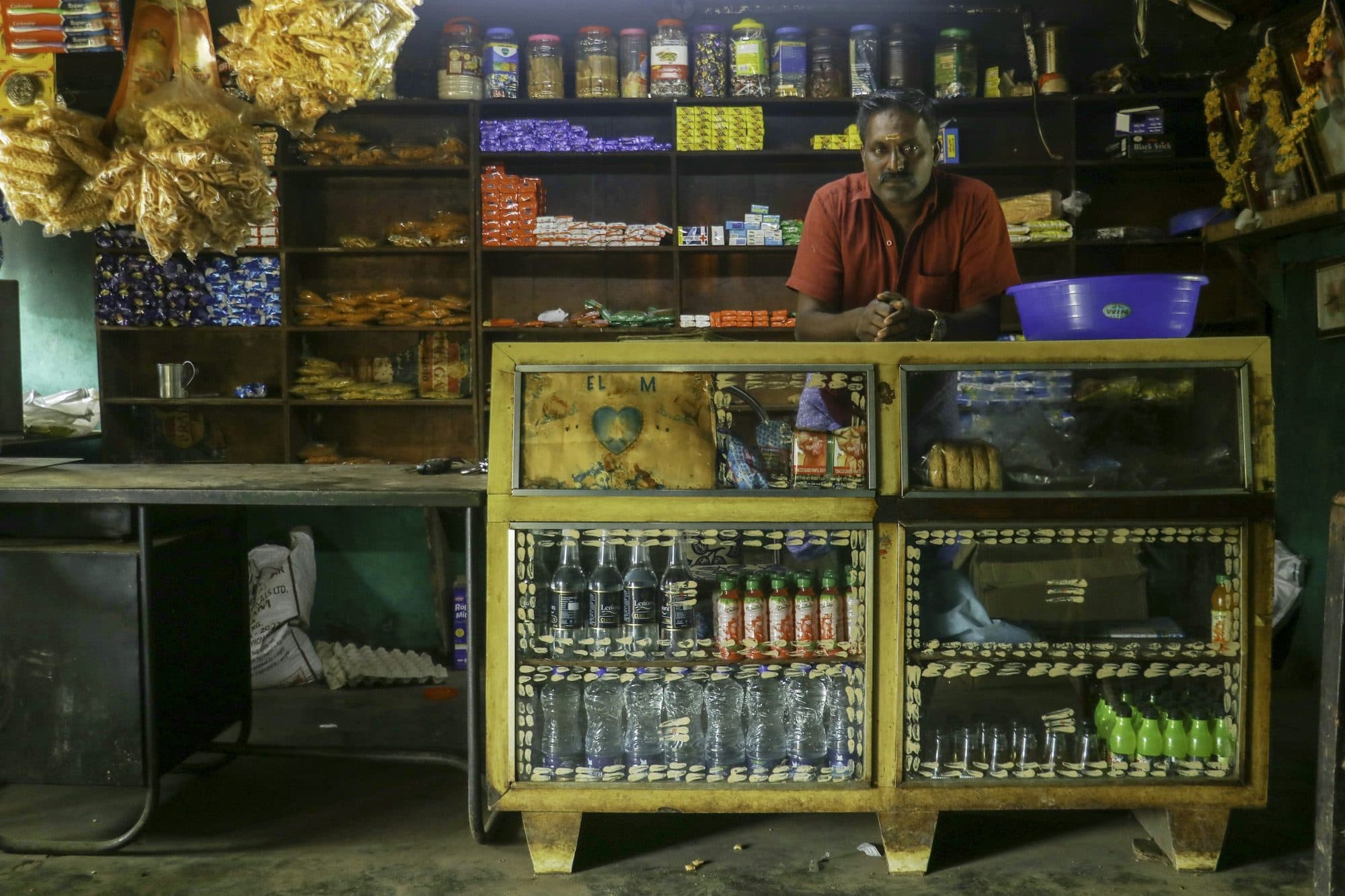 A shopkeeper waiting behind the counter of a small grocery, stock shelved to the ceiling behind him