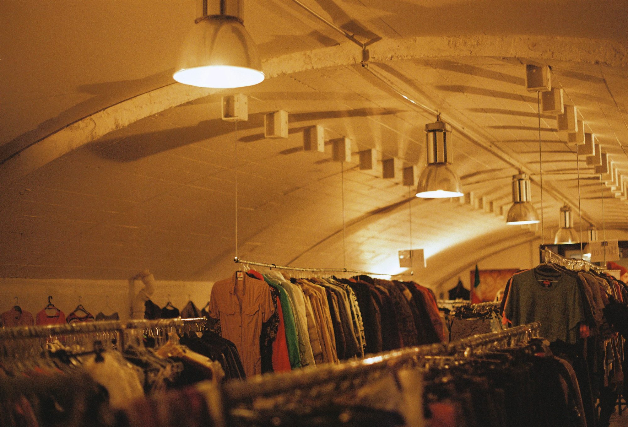 Rails of second-hand clothing running the length of a vaulted brick cellar under hanging lamps